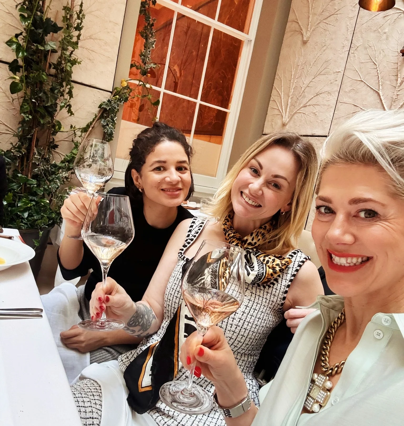 Three women raise glasses of rosé wine at a restaurant table, with plates and cutlery visible.