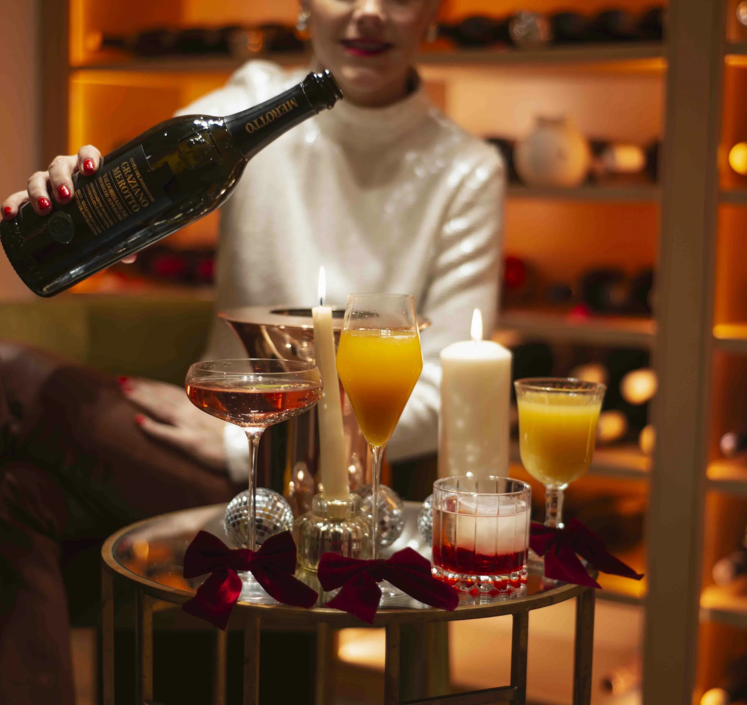 Person pouring Prosecco beside Christmas cocktails, candles and red bows