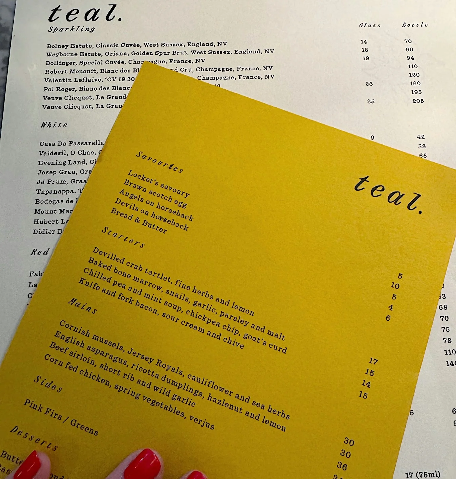 Teal restaurant menu with sparkling wines and dishes, partly held by a hand with red-painted nails