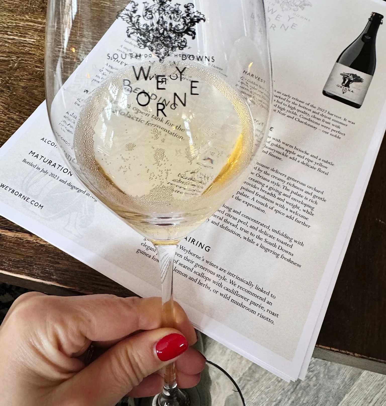 Hand holding a glass of sparkling wine over a Weyborne wine information sheet