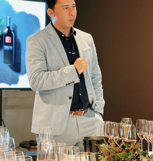 Wine presenter speaking beside a tasting table with glasses and a projected bottle