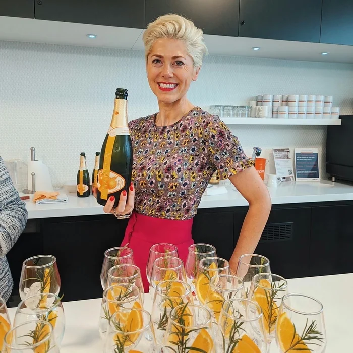 Elizabeth holds a bottle of sparkling wine behind glasses garnished with orange and rosemary at a tasting event