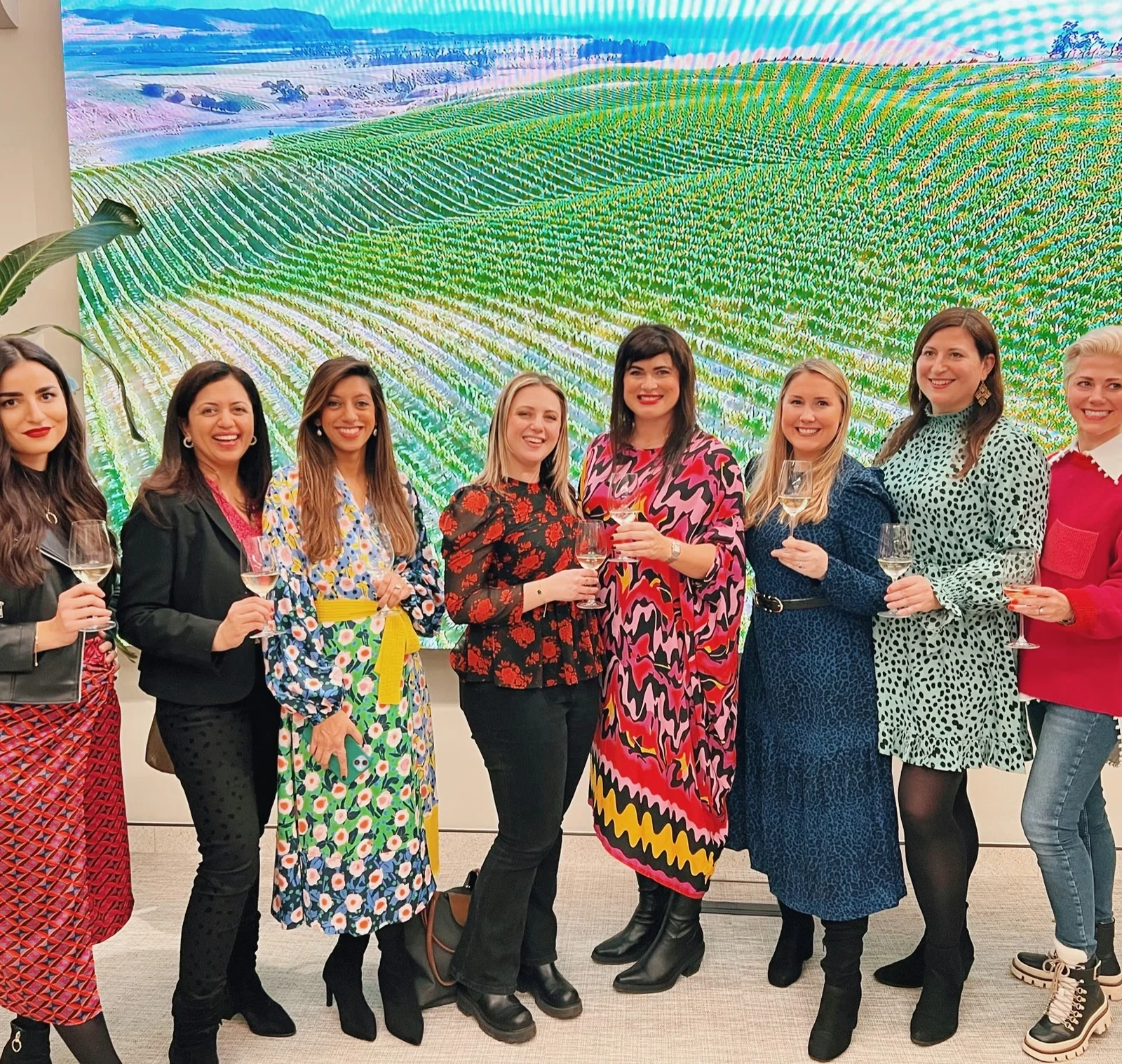 Eight women holding wine glasses at an International Women's Day event, standing before a vineyard display