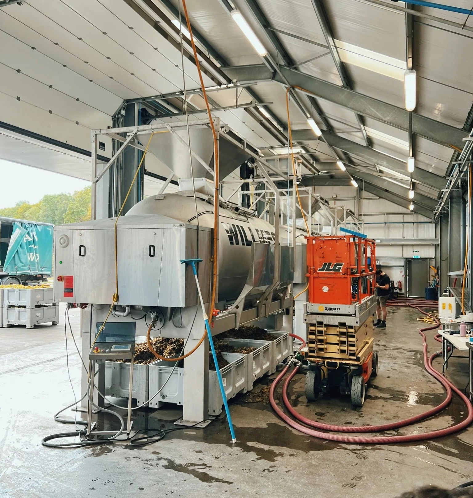 Winery interior with stainless steel tanks, equipment, hoses and a worker beside a red lift