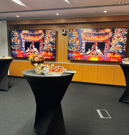 Catered wine event with a flower arrangement and canapés on a cocktail table, beside screens showing festive fireplaces.
