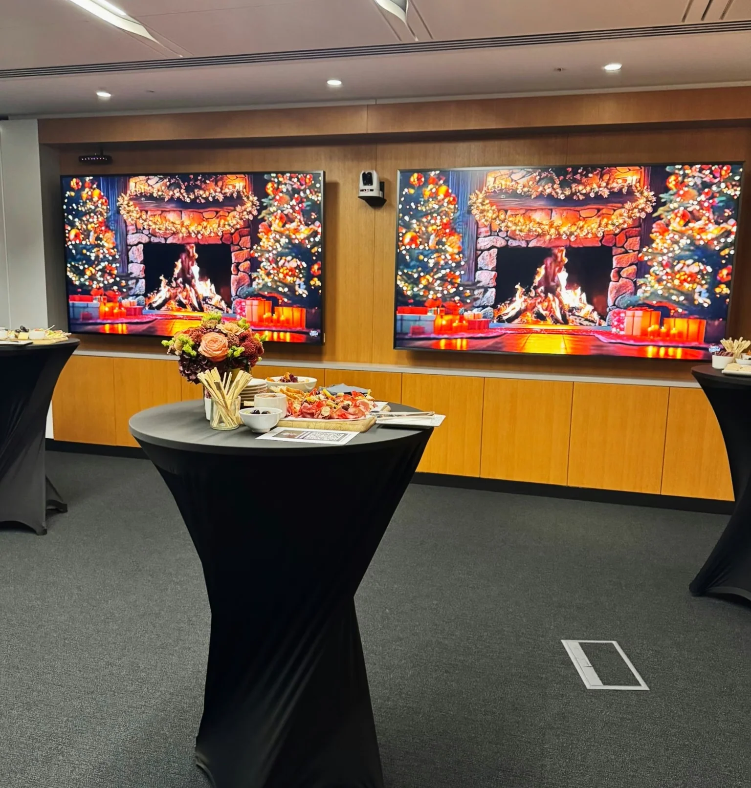 Catered wine event with a flower arrangement and canapés on a cocktail table, beside screens showing festive fireplaces.