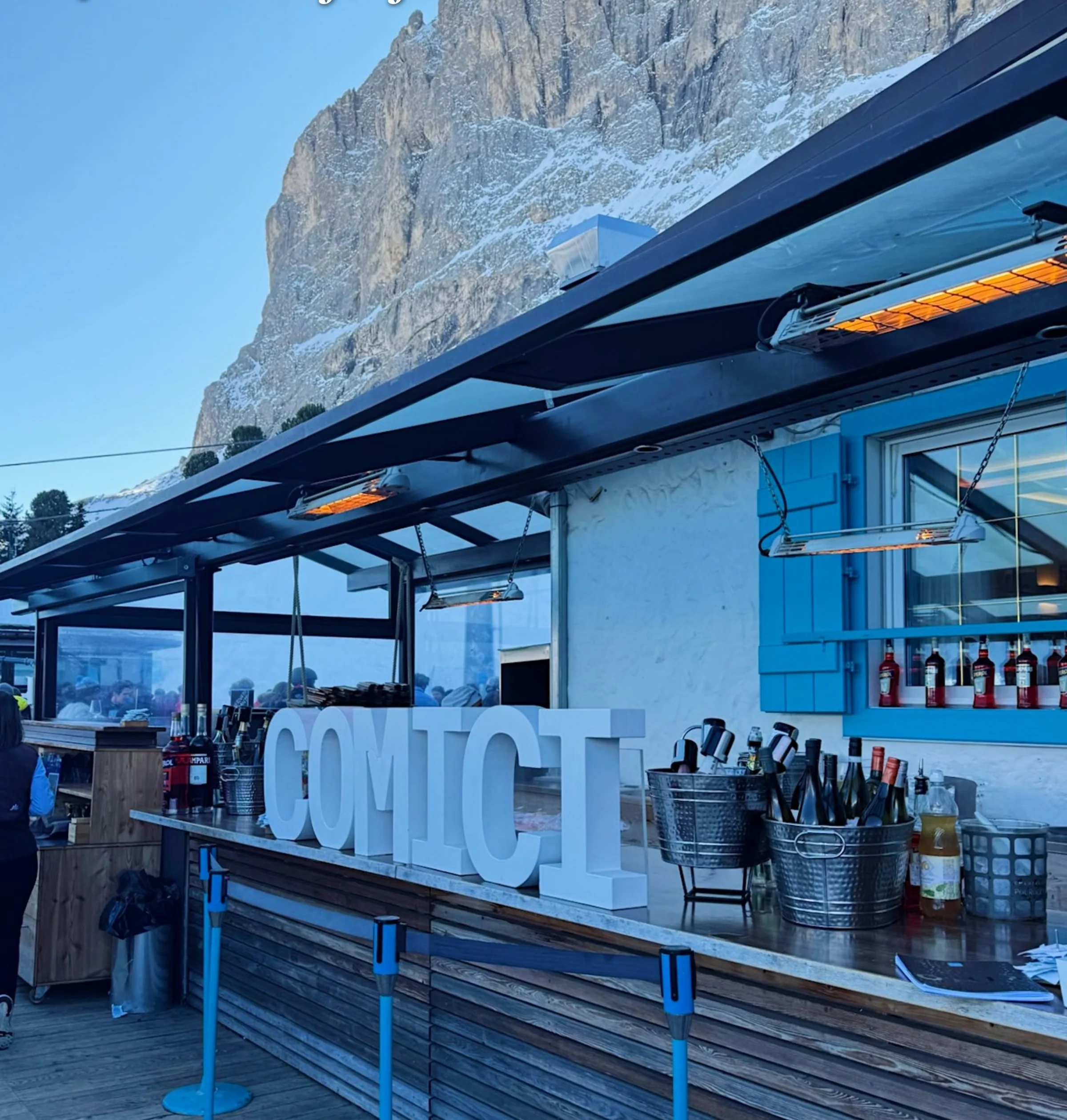 Outdoor restaurant bar with large COMICI letters beneath a snow-dusted Dolomite cliff
