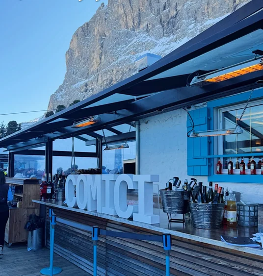 Outdoor restaurant bar with large COMICI letters beneath a snow-dusted Dolomite cliff