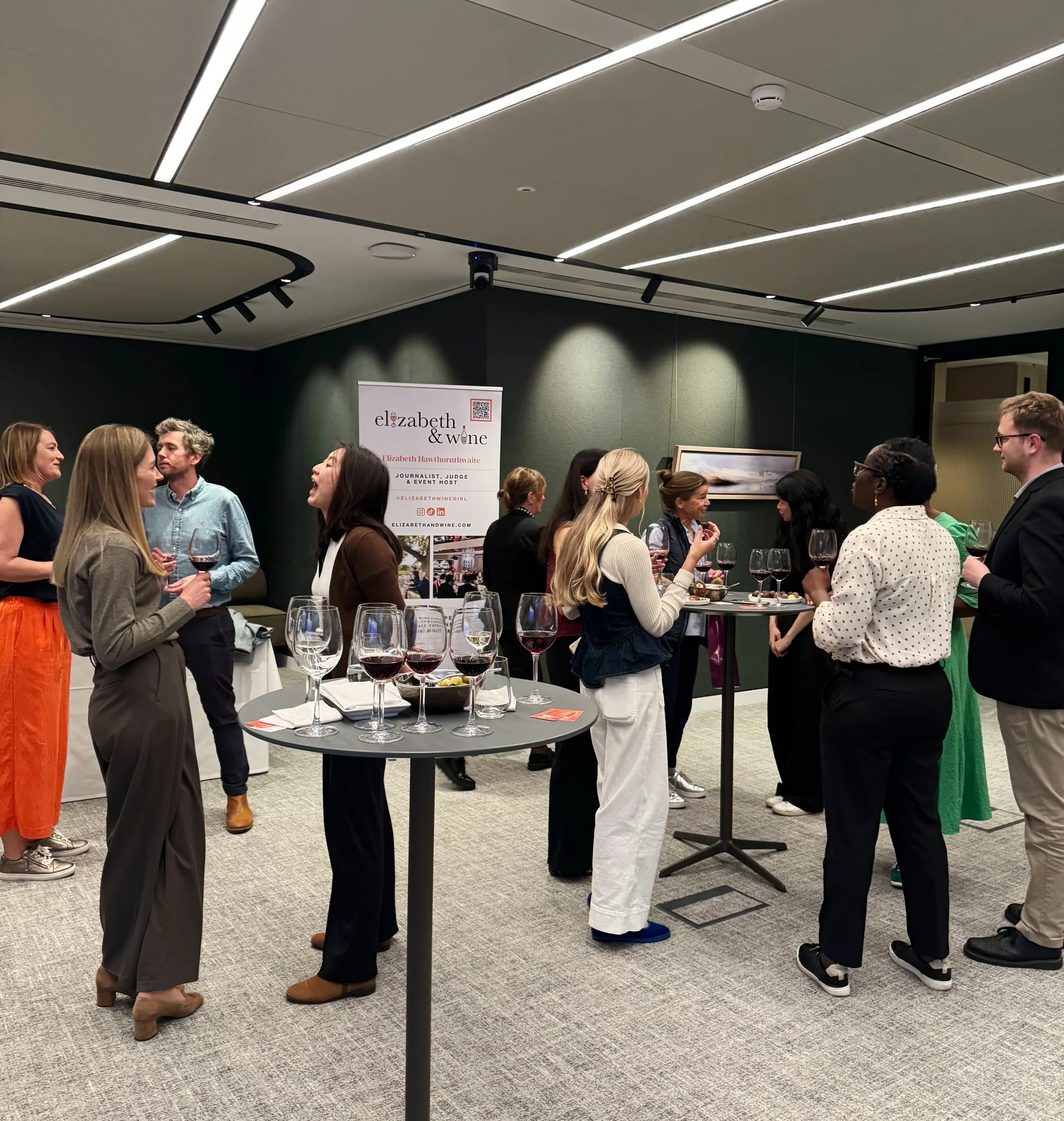 Guests mingle around wine tables at a networking event, with wine glasses and an Elizabeth & Wine banner.