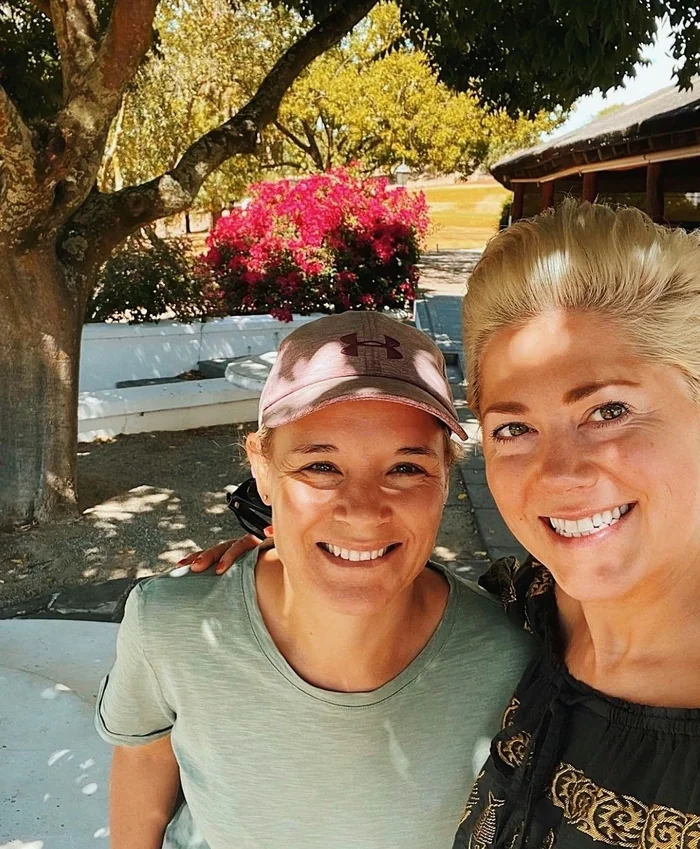 Two smiling women pose for a selfie outdoors beneath trees, with bright pink flowers behind them.