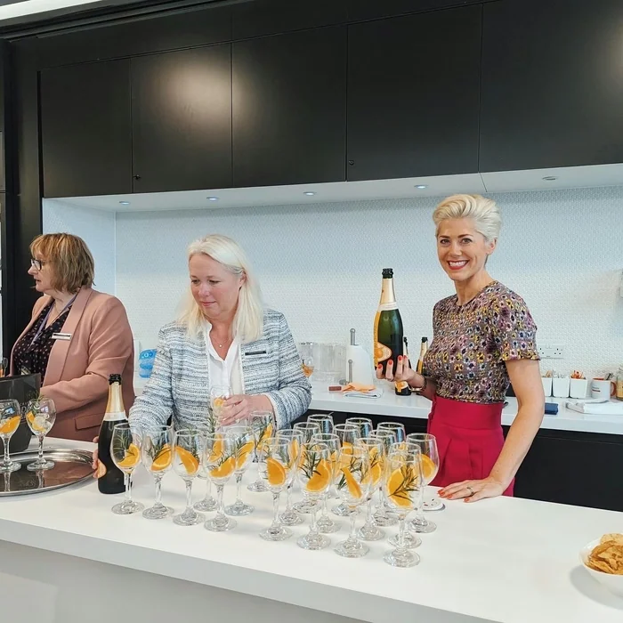 Three women serve wine at a tasting, with bottles and glasses garnished with orange slices and herbs.