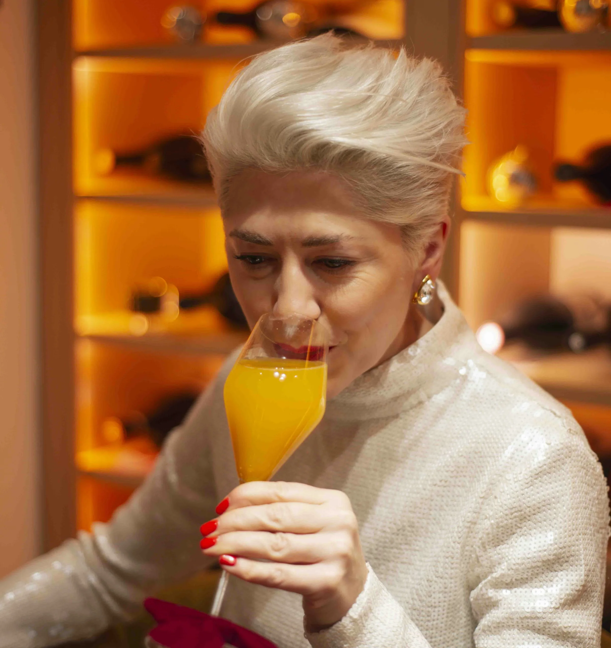 Person in a white jumper sips a bright orange cocktail in a warmly lit wine room