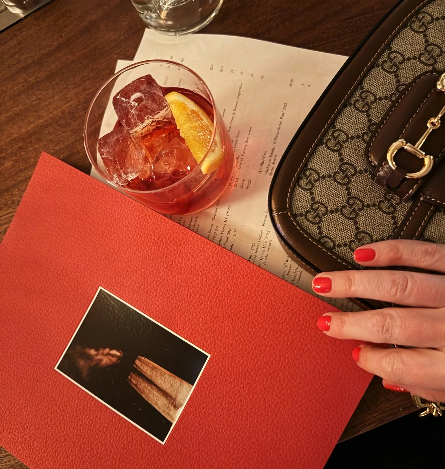 Red cocktail with ice and orange garnish beside a menu, red folder and patterned handbag