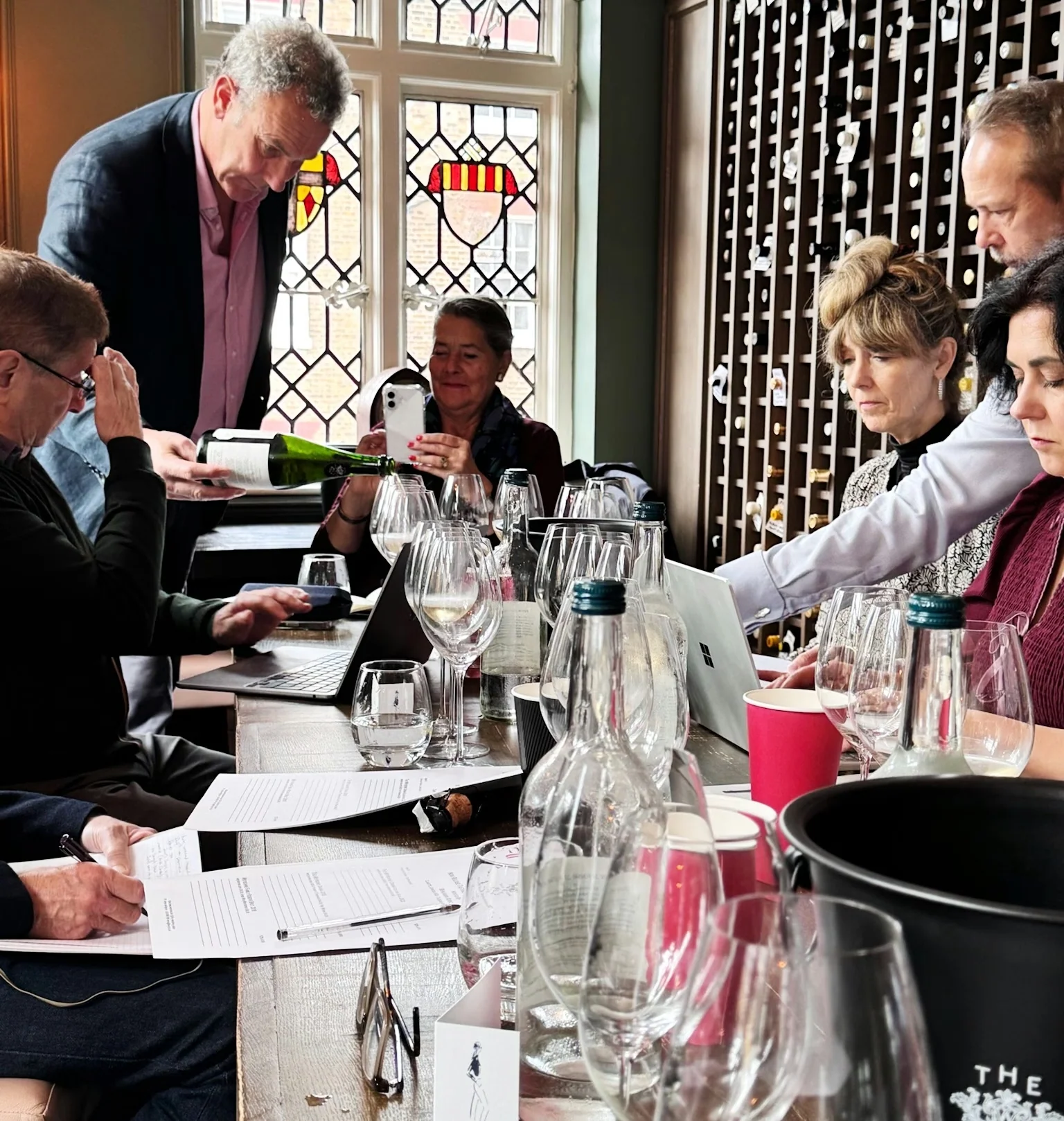 Guests taste wine around a table beside a wall of wine bottles