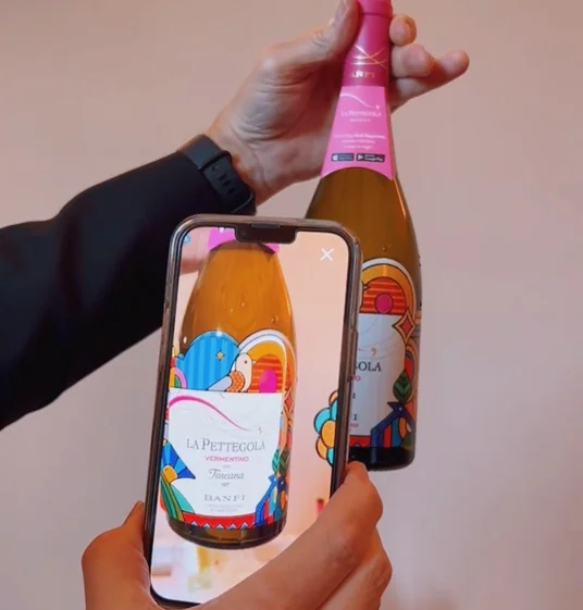 A hand holds a La Pettegola Vermentino bottle while another person photographs the label