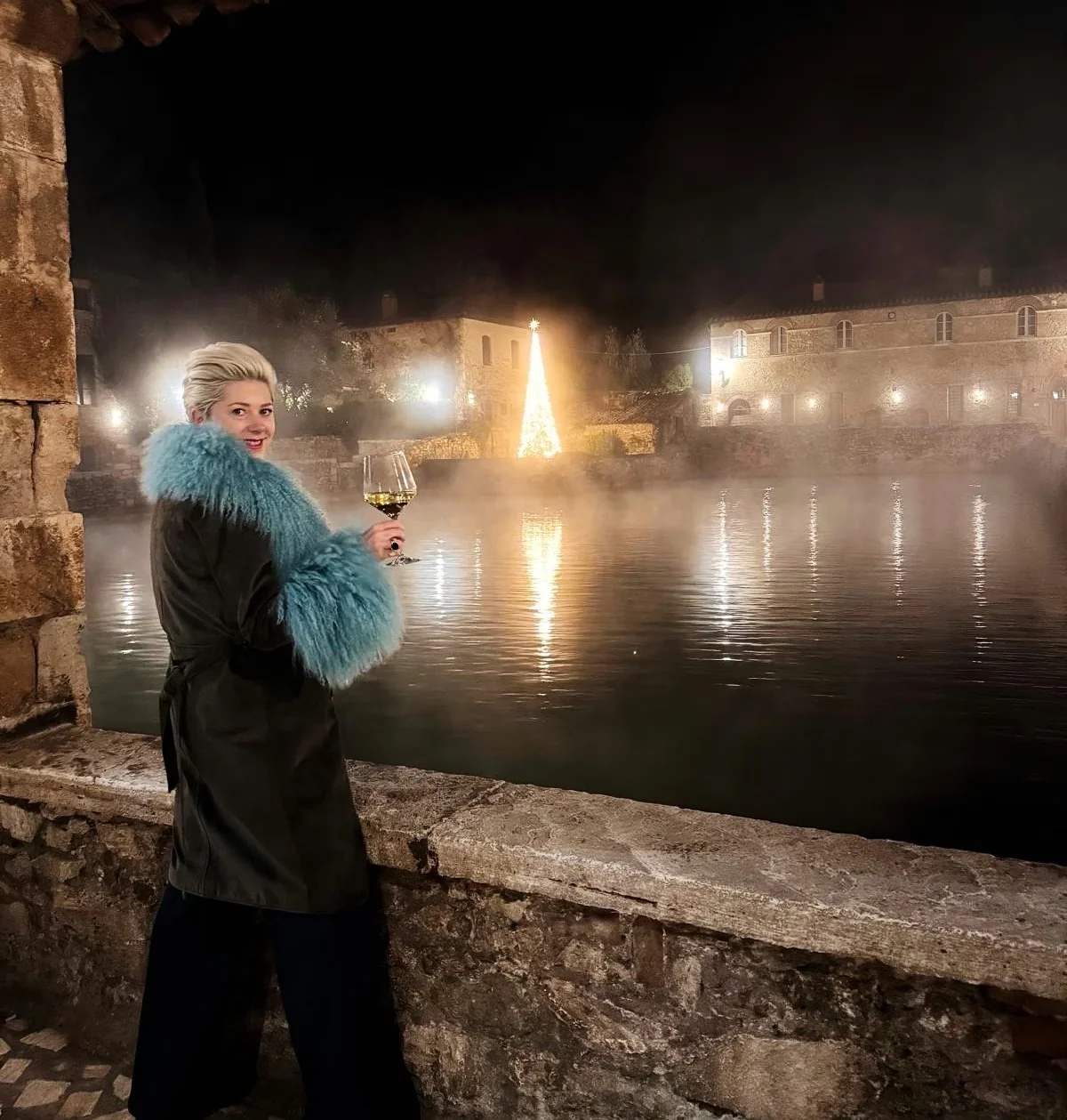 Person holding a wine glass beside a misty pool with a lit Christmas tree at night