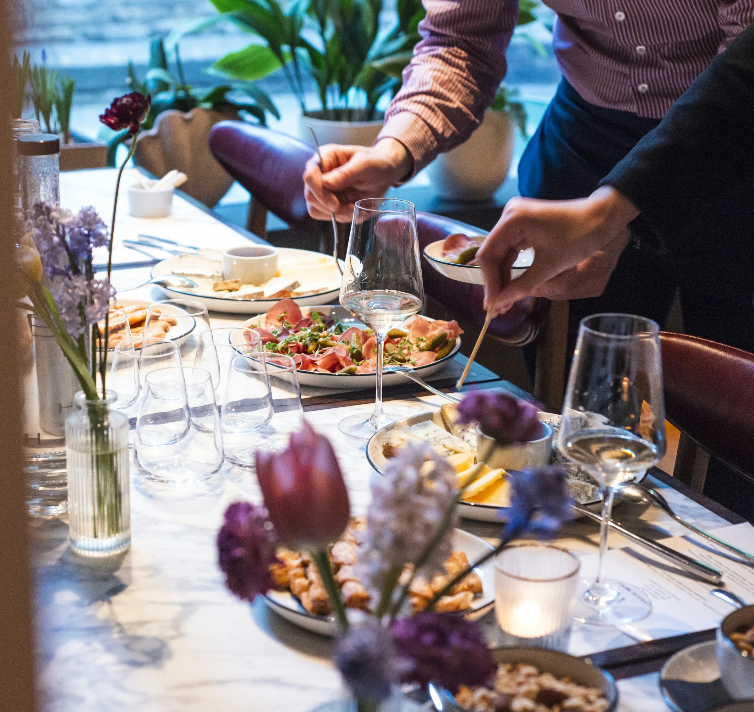 Chefs serve food at a dining table set with wine glasses, plates and flowers