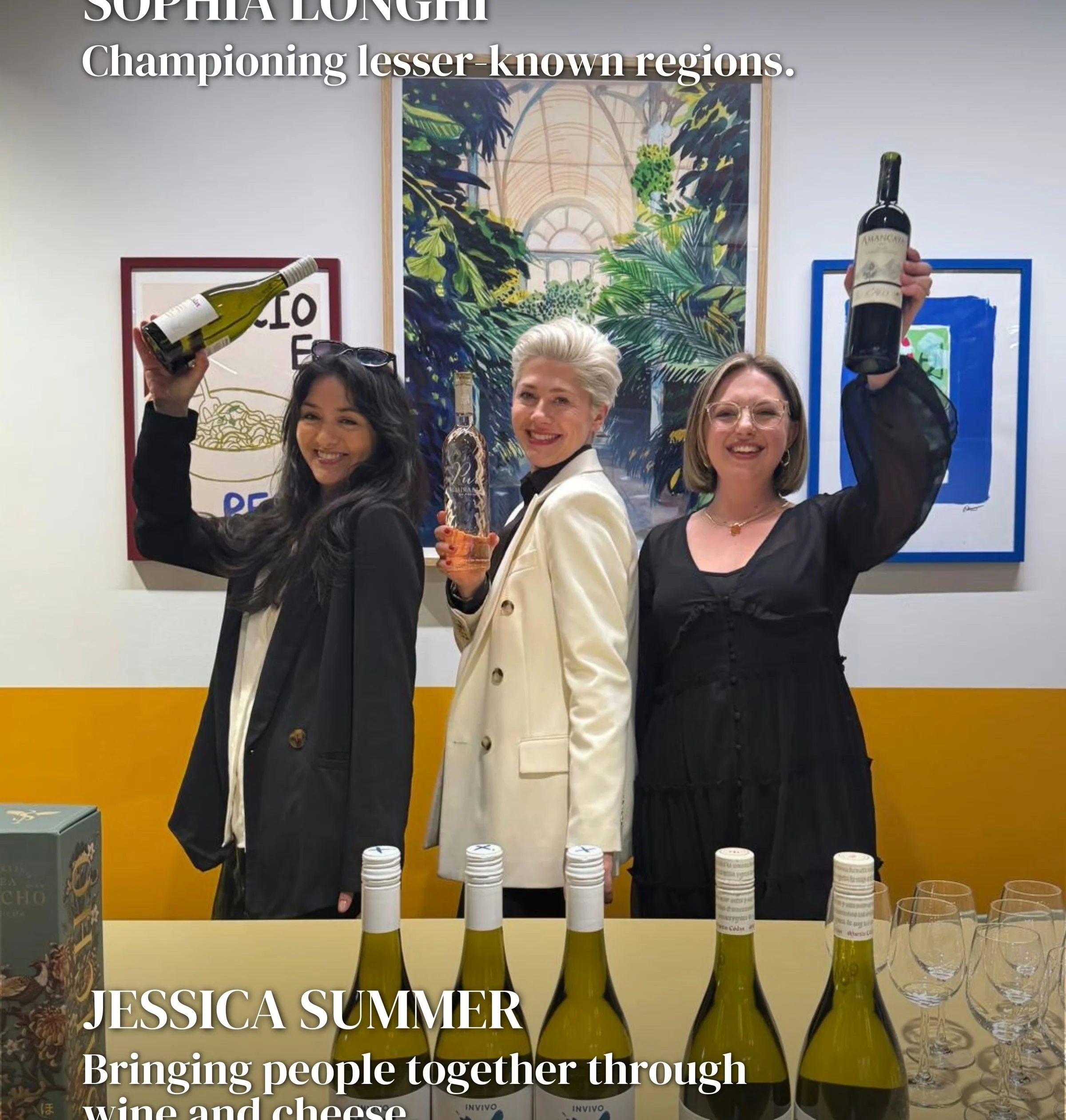 Three women raise wine bottles at a tasting table with wine glasses and bottles displayed in front