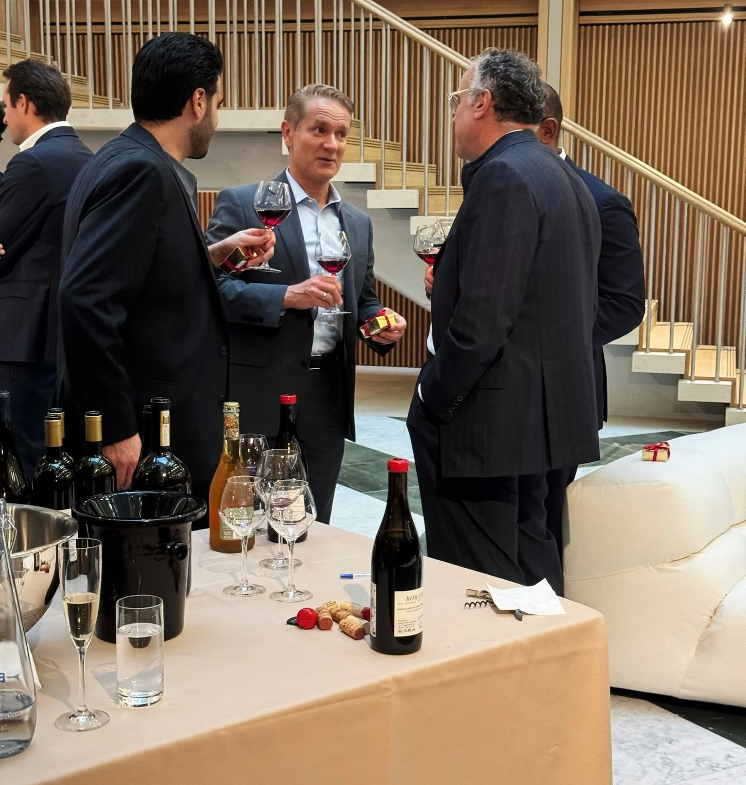 Colleagues chat over wine at an office tasting, beside a table of wine bottles and glasses