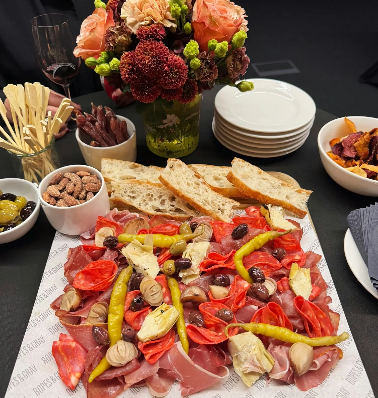 Catered wine tasting table with a floral arrangement, red wine, stacked plates, bread, nuts and a charcuterie board