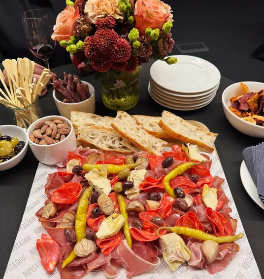 Catered wine tasting table with a floral arrangement, red wine, stacked plates, bread, nuts and a charcuterie board