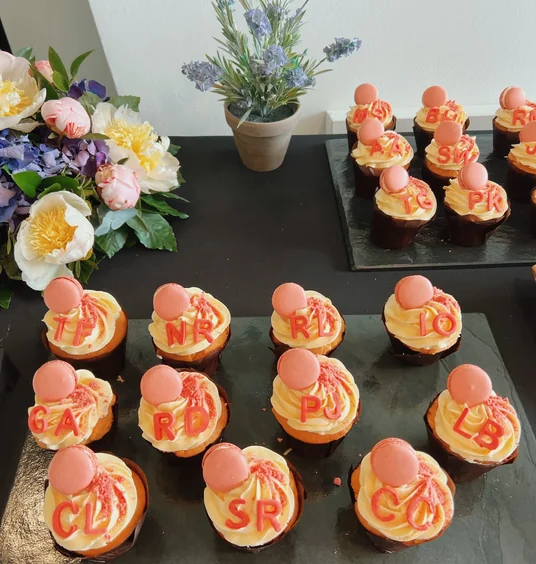 Decorated cupcakes with pink macarons and red letter toppers beside flowers and a potted plant