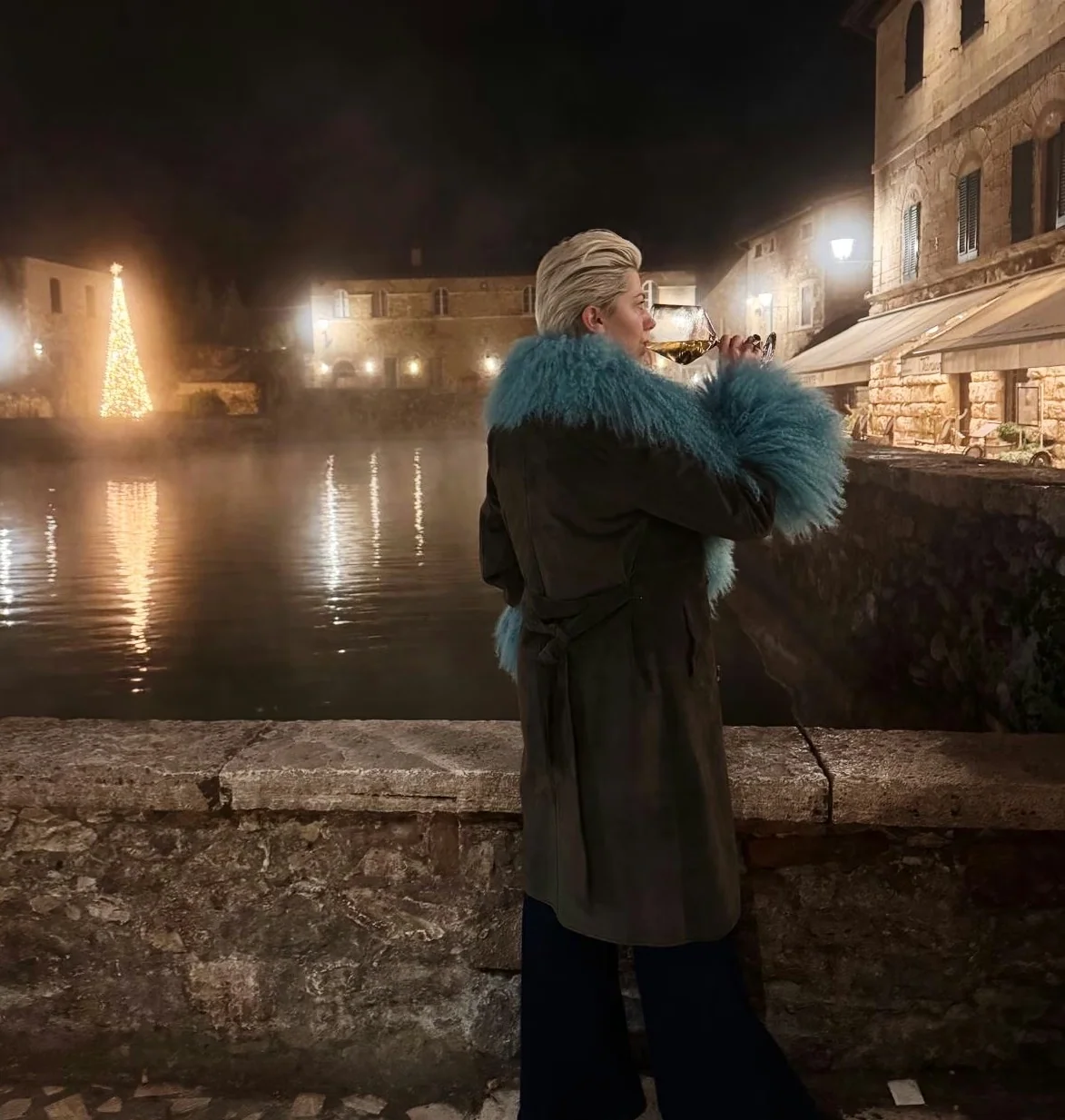Person tasting white wine beside a historic stone pool at night in Tuscany