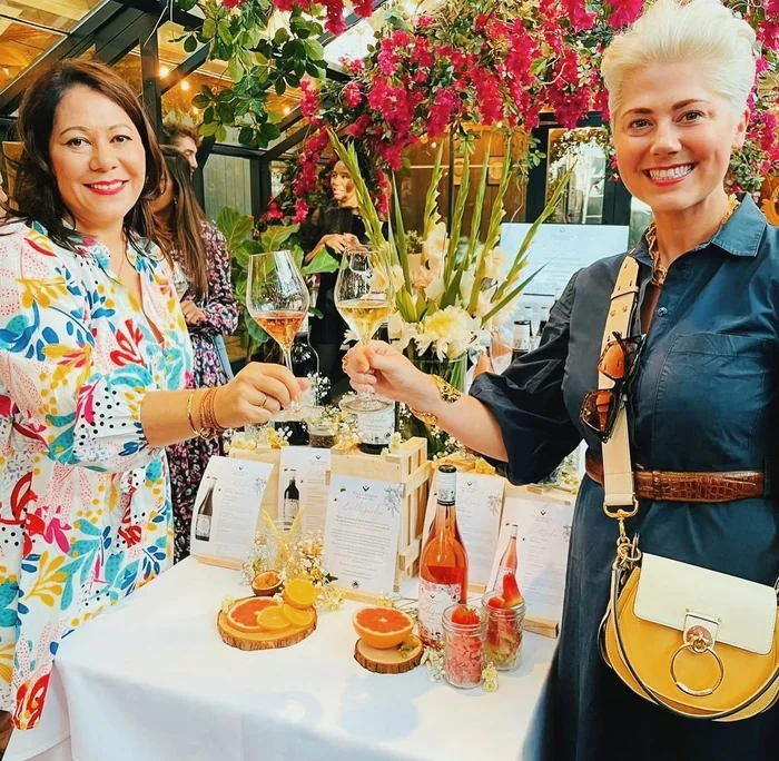 Two women toast with wine glasses at a colourful floral wine tasting display.