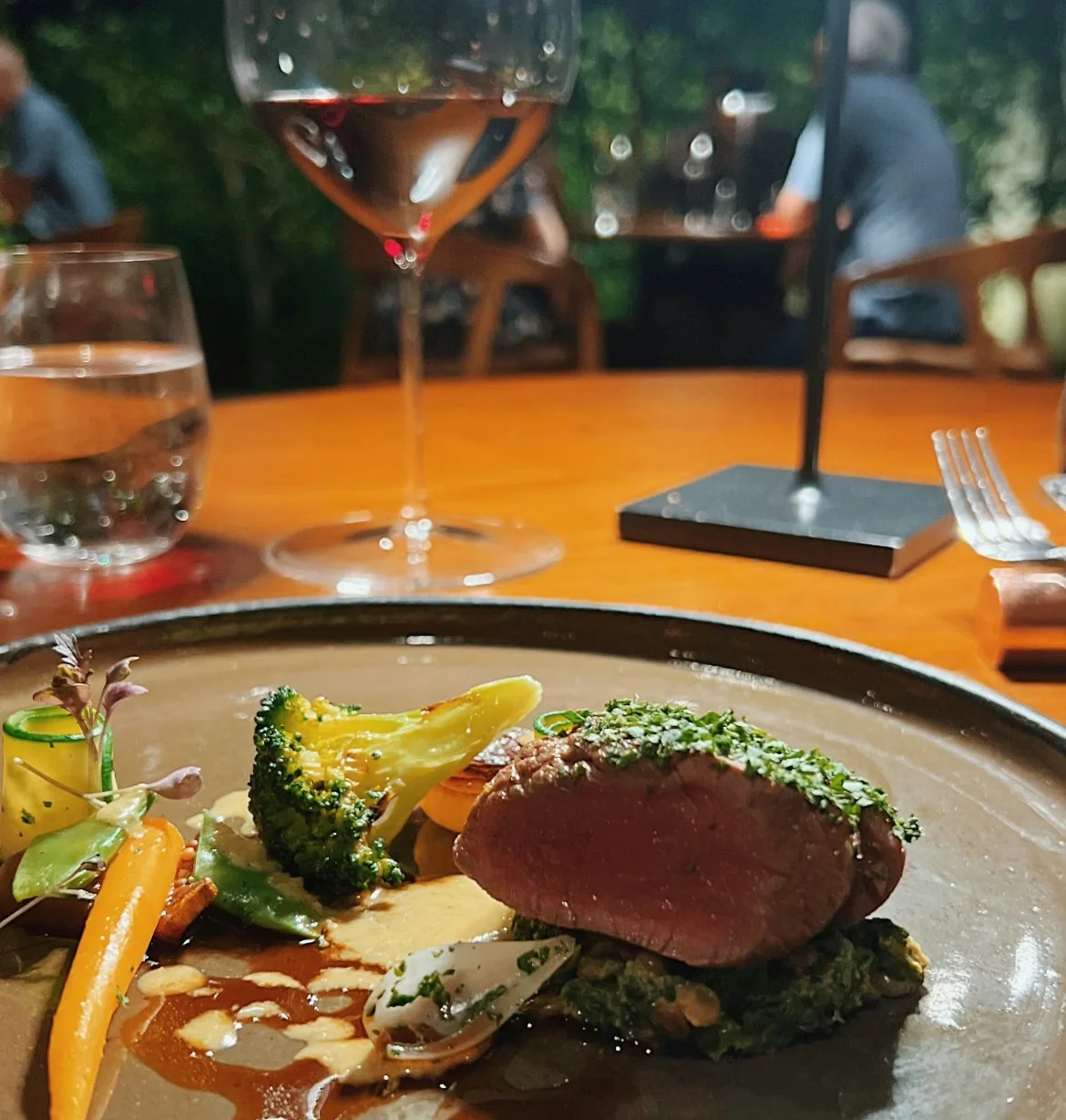Restaurant table with a plate of steak, vegetables and sauce beside a glass of red wine