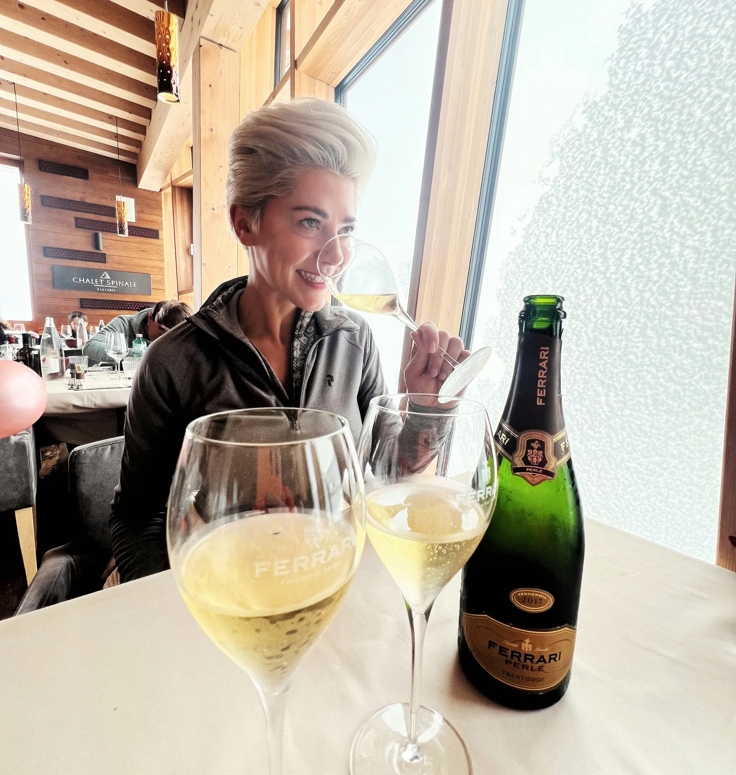 Woman tasting sparkling wine beside a Ferrari Perlé bottle in a wooden mountain restaurant