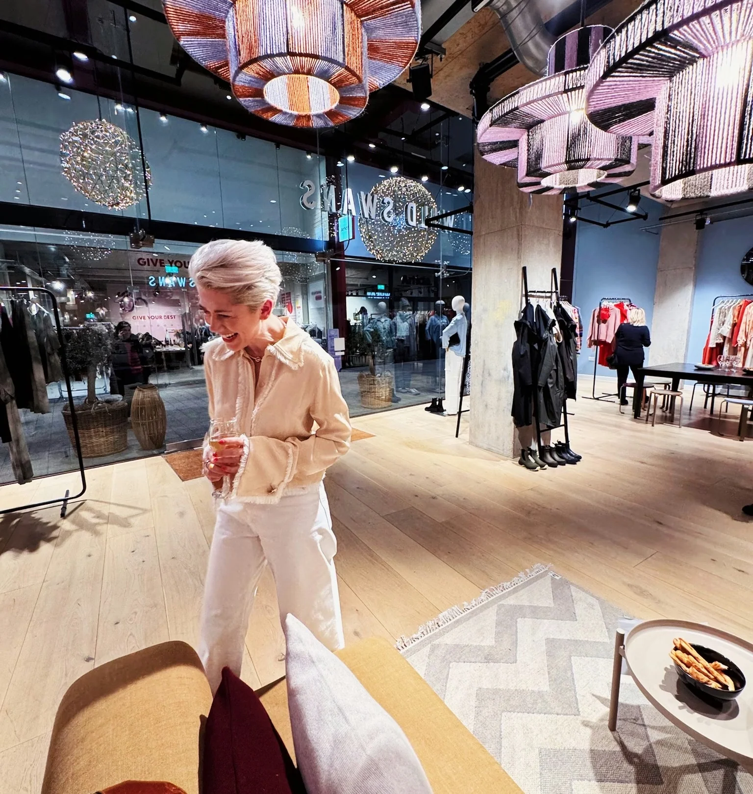 Woman holding a wine glass in a bright fashion shop beneath large illuminated pendant lights