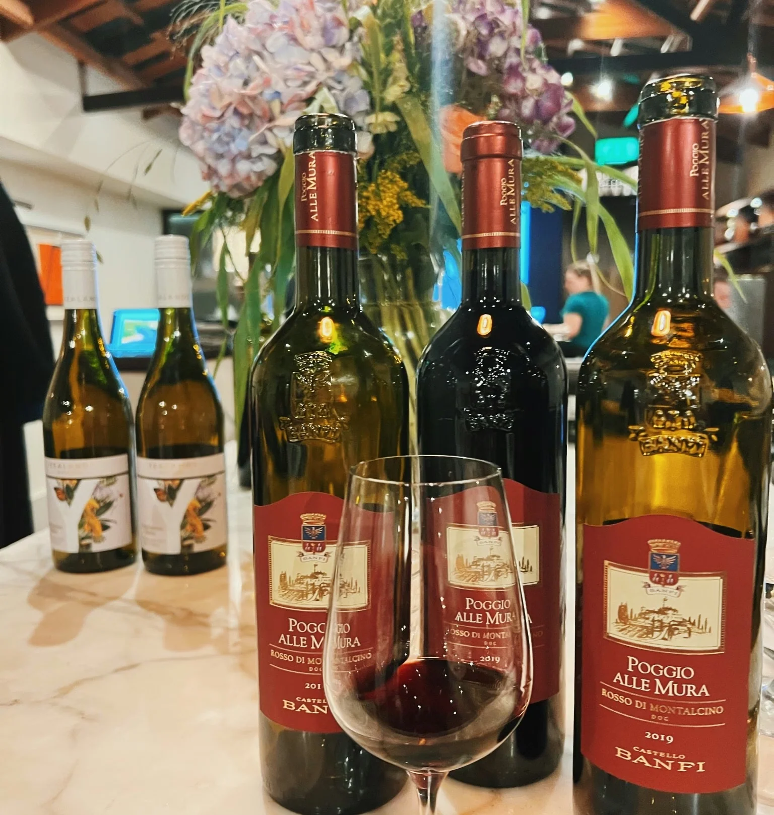 Poggio alle Mura wine bottles and a glass of red wine beside a floral arrangement