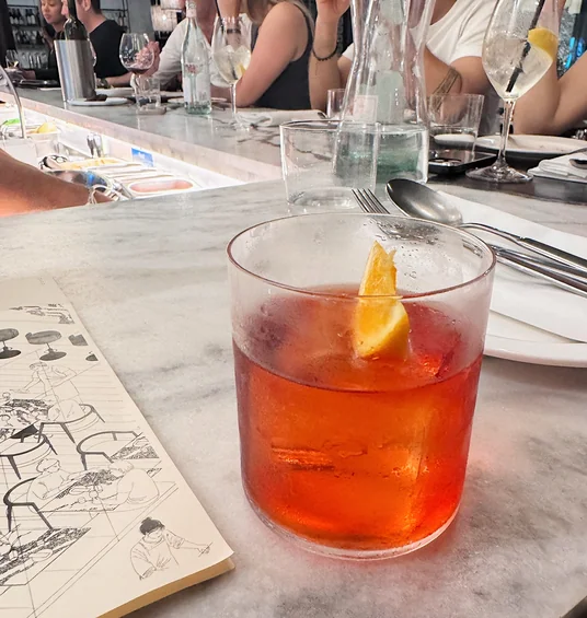 Red cocktail with an orange garnish on a restaurant counter, with diners and wine glasses behind