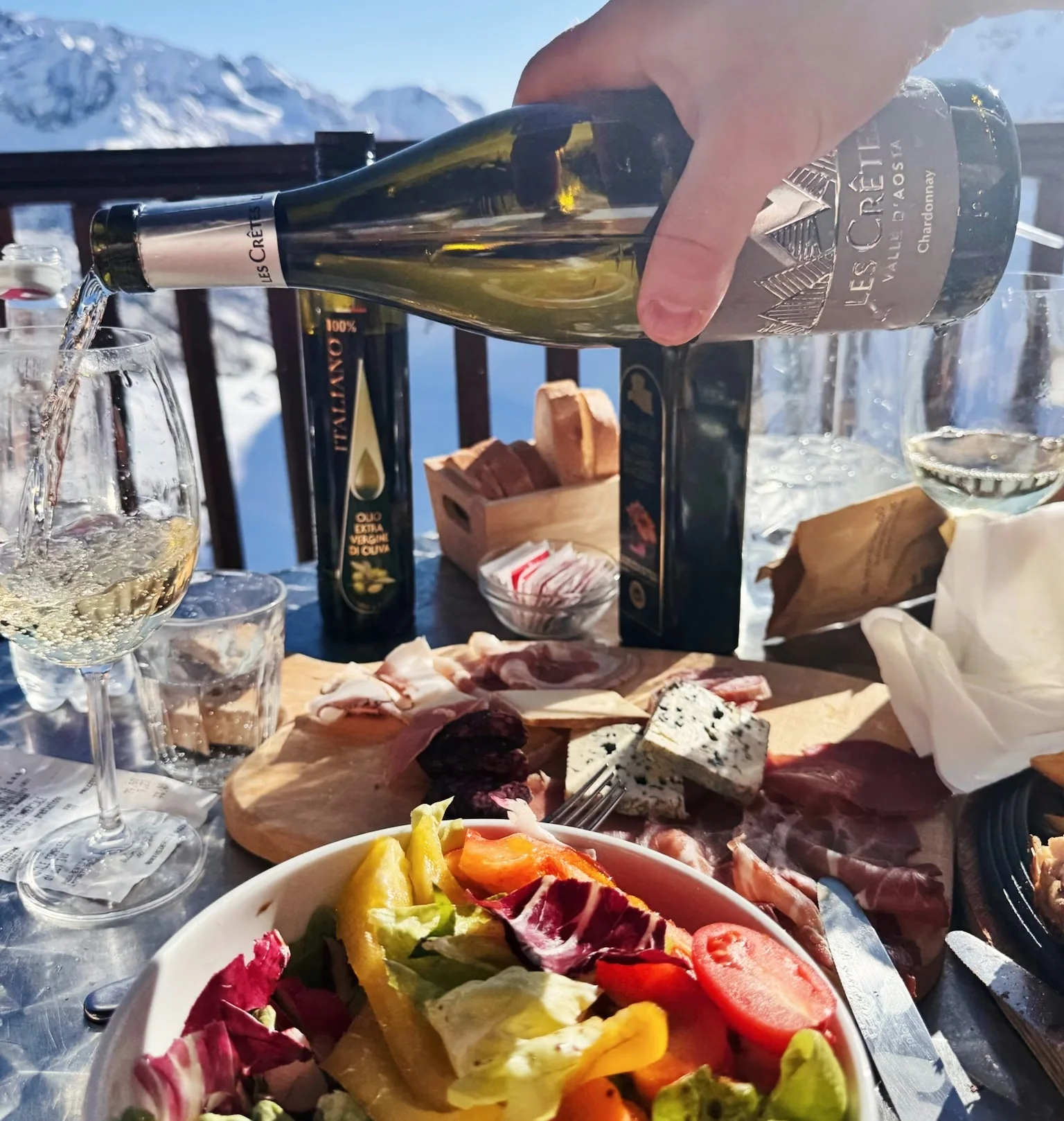 A hand pours Les Crêtes Chardonnay beside mountain views, charcuterie, cheese and salad.