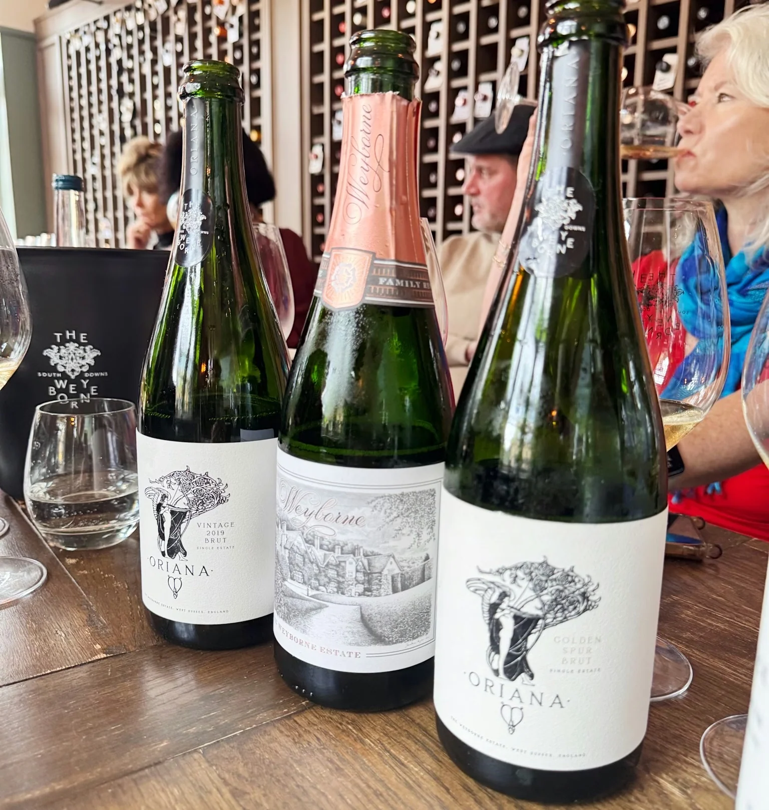 Three English sparkling wine bottles on a tasting table with guests and a bottle display behind