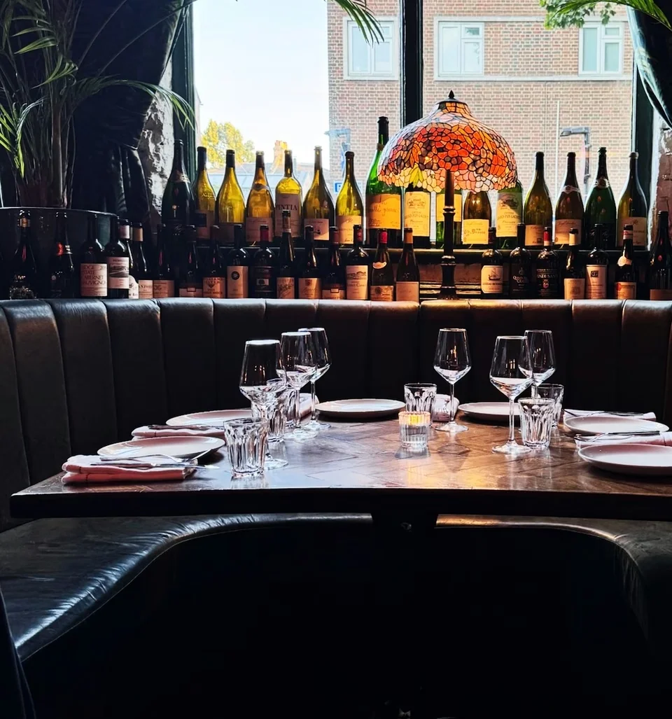 Restaurant table set with wine glasses beneath a display of wine bottles and a Tiffany-style lamp