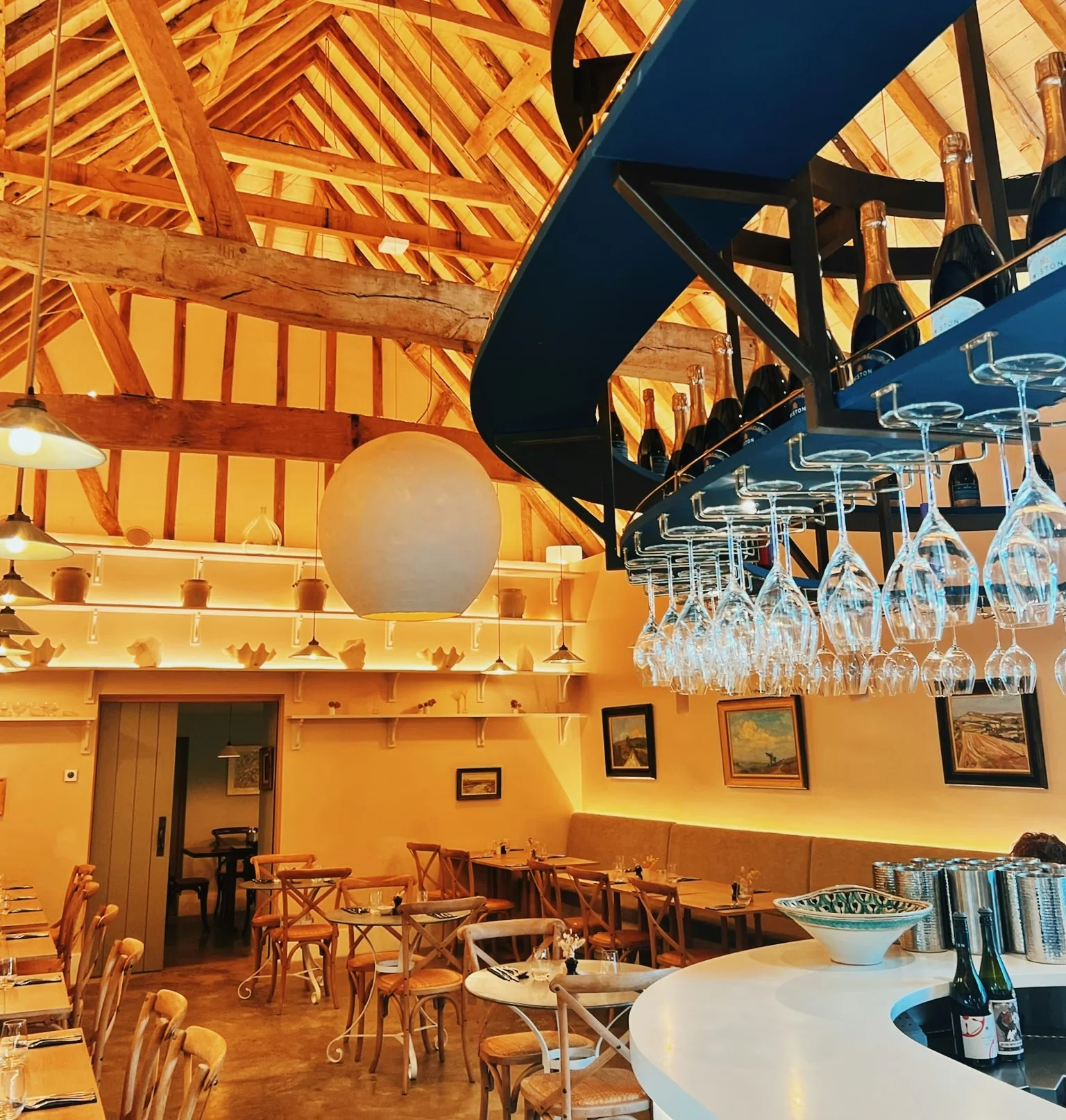 Warmly lit restaurant interior with timber beams, a curved bar, hanging glasses and bottles of sparkling wine