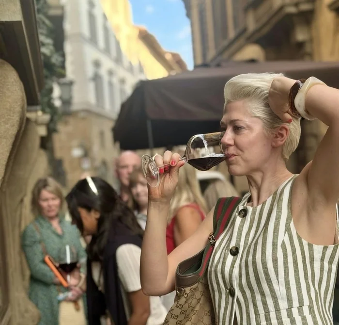 Woman drinks red wine from a glass beside a historic wine window in a Florence street.