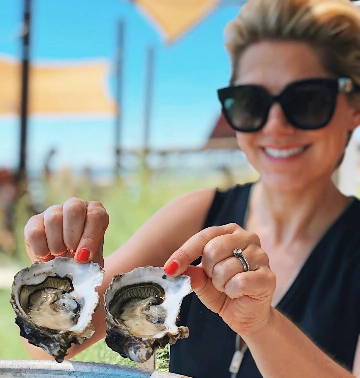 Smiling woman in sunglasses holds two freshly opened oysters at an outdoor tasting