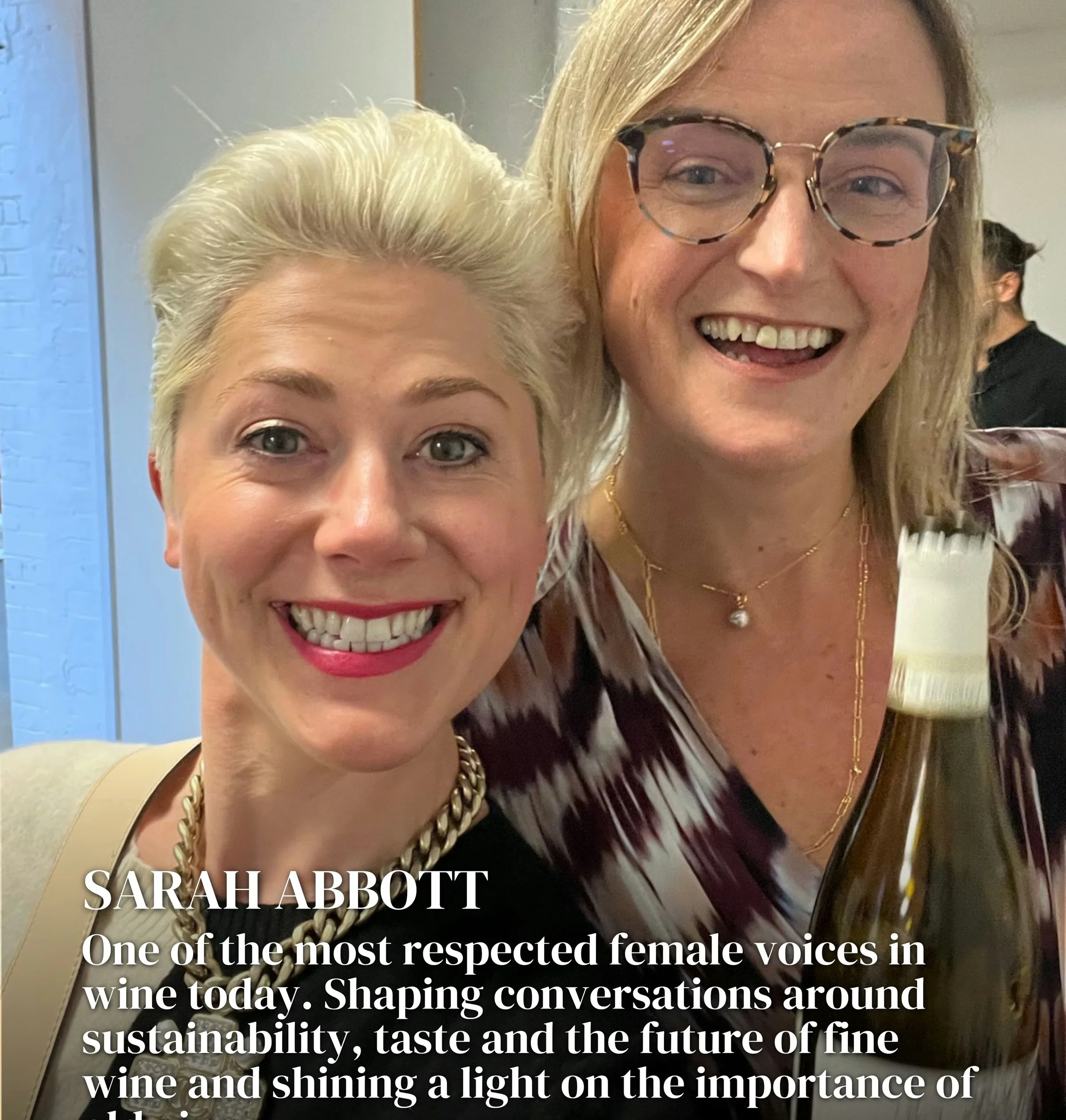 Two smiling women pose for a selfie, with a wine bottle and text about Sarah Abbott in the foreground