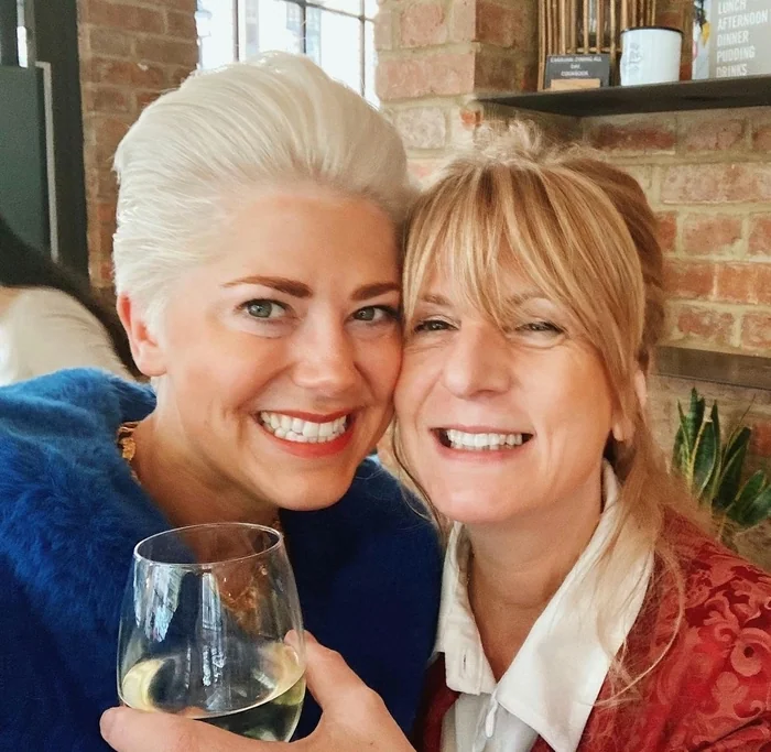 Two women smiling together in a brick-walled venue, with a glass of white wine in the foreground