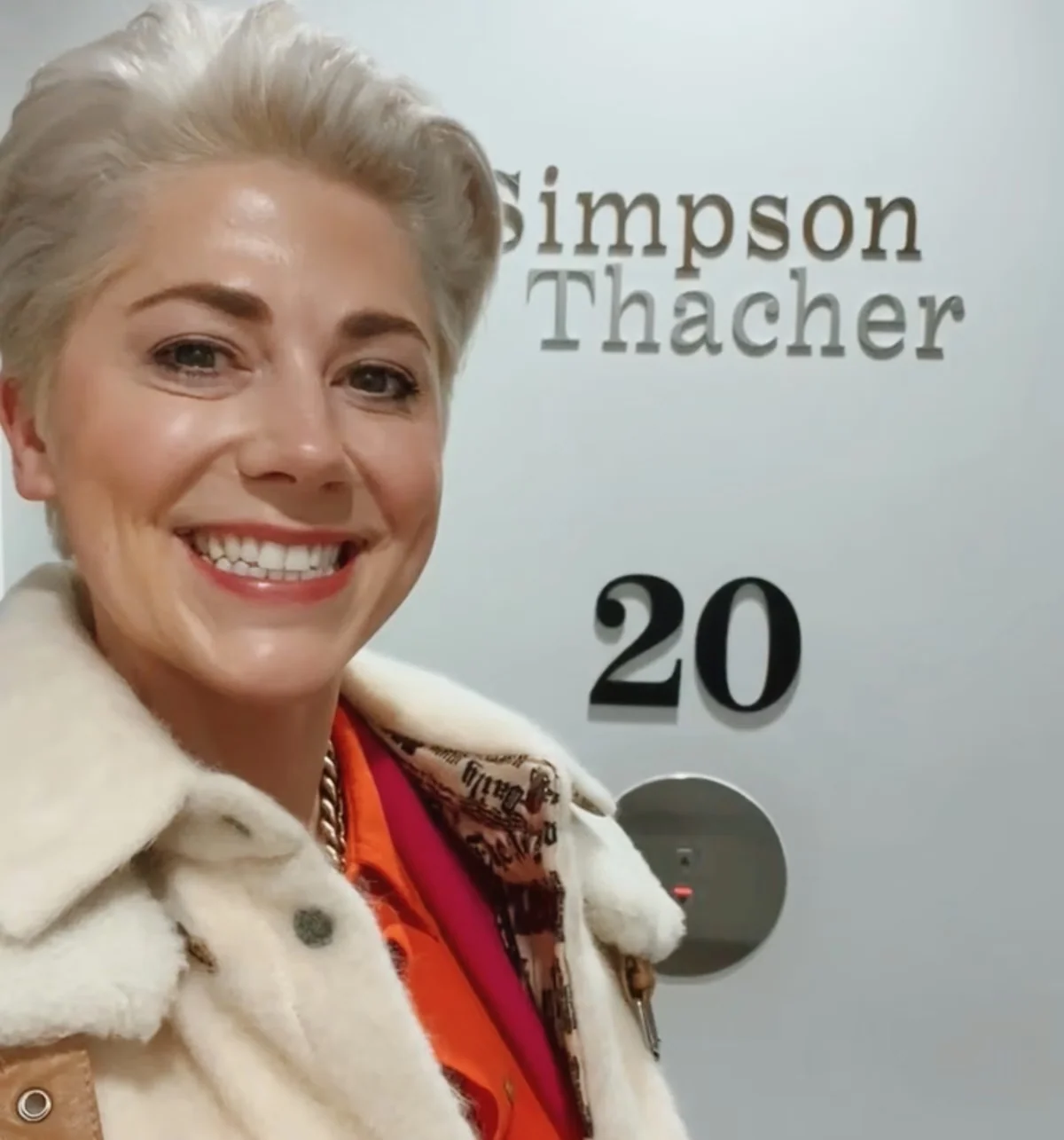 Smiling woman in a white coat and colourful scarf beside Simpson Thacher signage