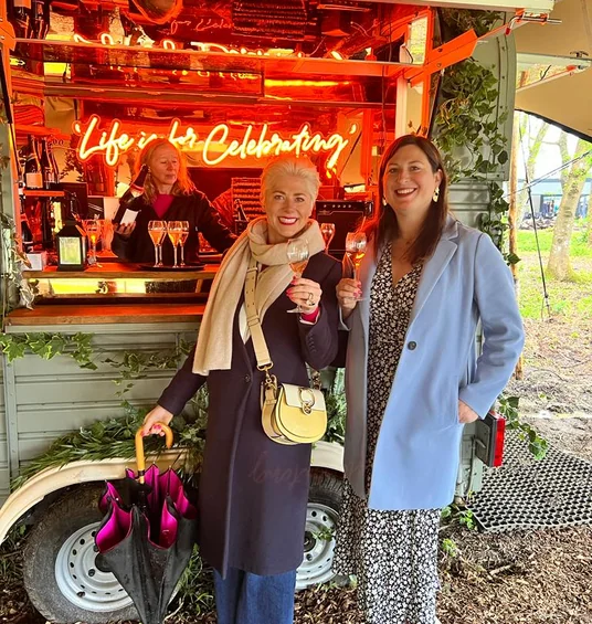 Two women raise wine glasses beside a brightly lit mobile bar with a neon celebration sign
