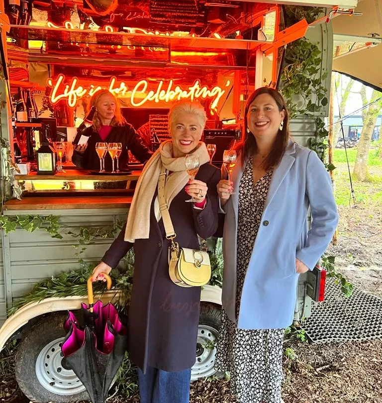 Two women raise wine glasses beside a brightly lit mobile bar with a neon celebration sign