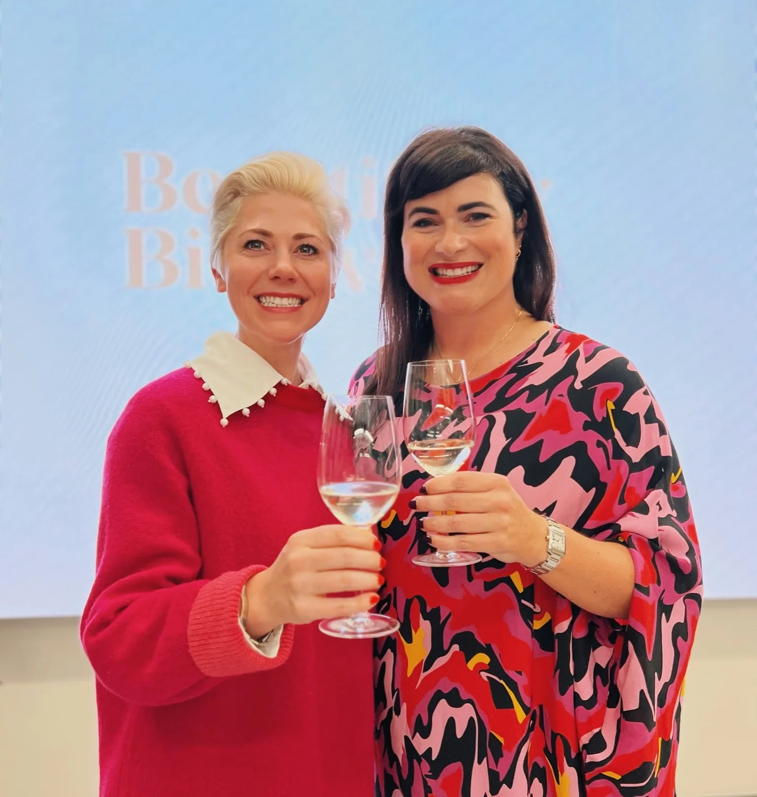 Two women in colourful clothing smile and raise wine glasses at an International Women's Day event