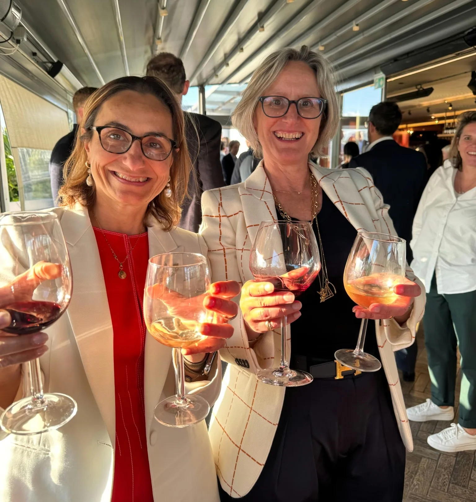 Two women smile and hold glasses of wine at a busy networking event.