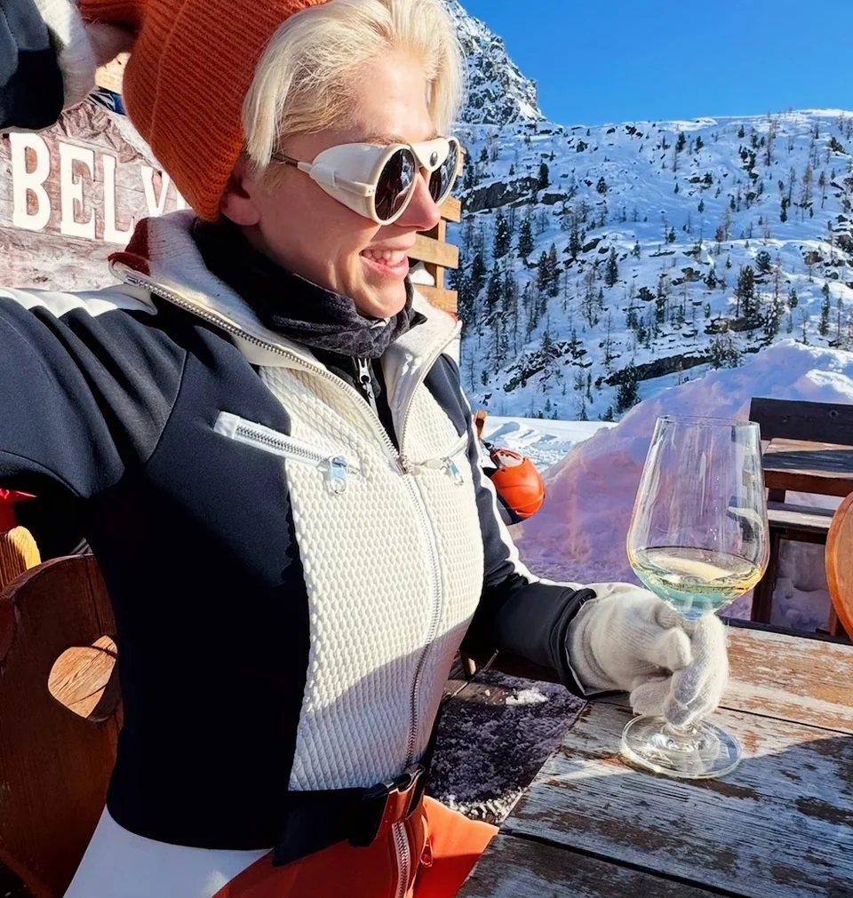 Skier in sunglasses and orange hat holding a glass of white wine on a snowy terrace in Champoluc