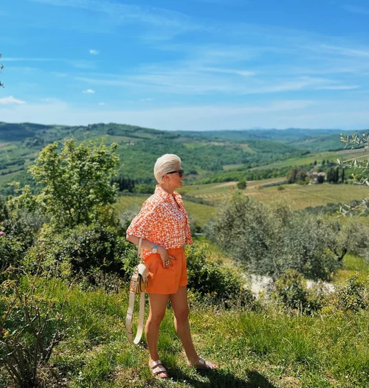 Woman in orange clothing standing among vines, with rolling Tuscan hills and vineyards behind her