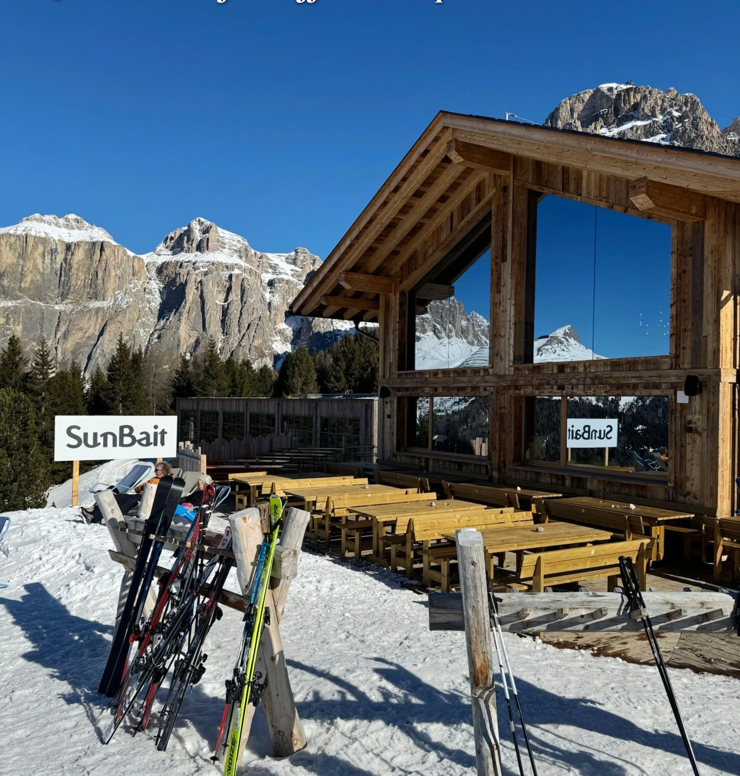 Sun Bait mountain restaurant with outdoor benches, skis and snow-covered Dolomite peaks