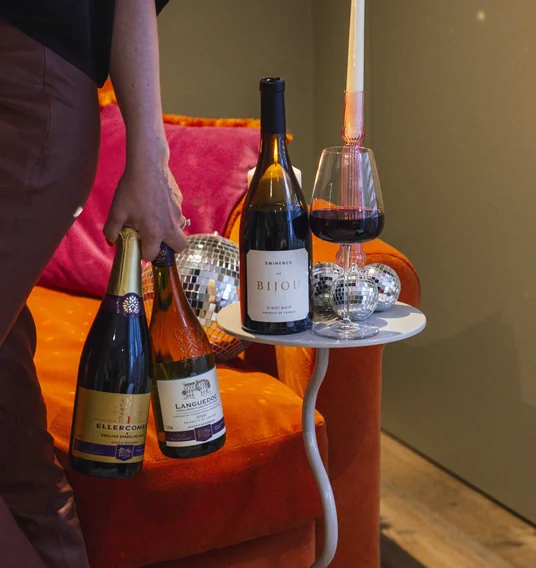 Wine bottles and a red wine glass beside a lit candle in a colourful lounge setting